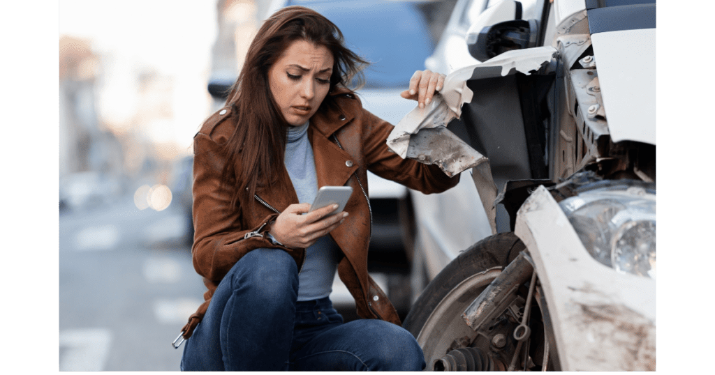 A woman sits beside her wrecked car, intently looking at her phone for assistance or information.